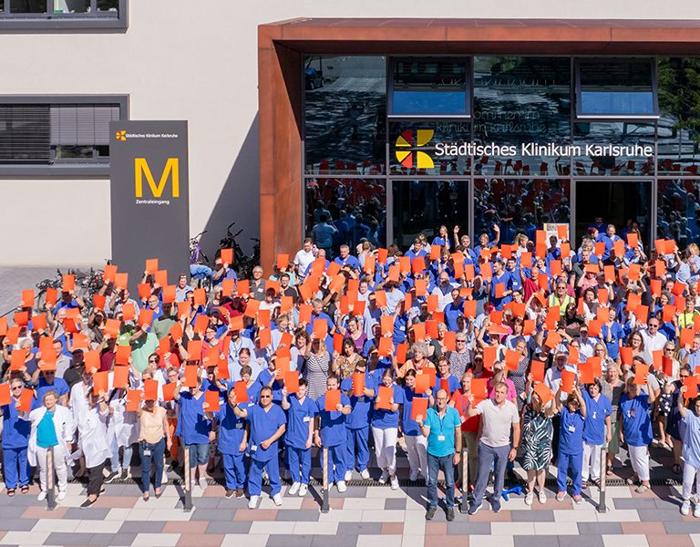 Das Bild zeigt ein Gruppenbild im Rahmen der Kampagne "Alarmstufe ROT". Mitarbeitende des Klinikums heben vor dem Eingang von Haus M stehend symbolisch eine Rote Karte in die Luft.