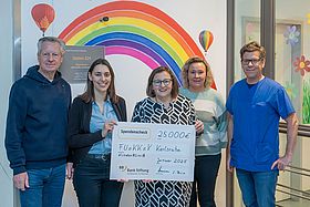 Das Bild zeigt sechs Personen bei der Übergabe eines Spendenchecks. Die Personen stehen am Eingang der Kinderonkologischen Station "Regenbogen", um Hintergrund ist auf der Wand ein großer, bunter Regenbogen aufgemalt.