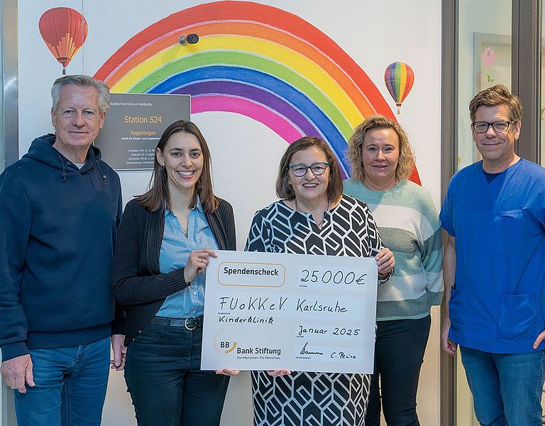 Das Bild zeigt sechs Personen bei der Übergabe eines Spendenchecks. Die Personen stehen am Eingang der Kinderonkologischen Station "Regenbogen", um Hintergrund ist auf der Wand ein großer, bunter Regenbogen aufgemalt.