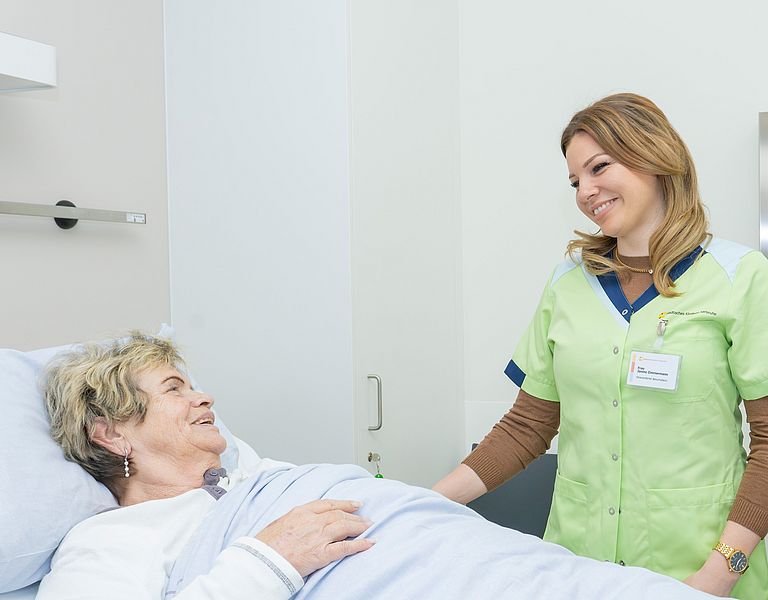 Zwei Mitglieder des ehrenamtlichen Begleitdienstes sprechen mit einer Patientin, die im Bett liegt.