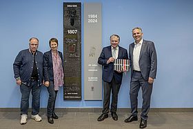Das Bild zeigt Künstler Ilko Uhde, Bürgermeisterin Bettina Lisbach, Prof. Martin Storck und Geschäftsführer MArkus Heming vor dem Kunstwerk. Das Kunstwerk besteht aus zwei Stelen, die an einer blauen Wand hängen. Auf einer Stele ist ein Bambi-Kopf montiert.
