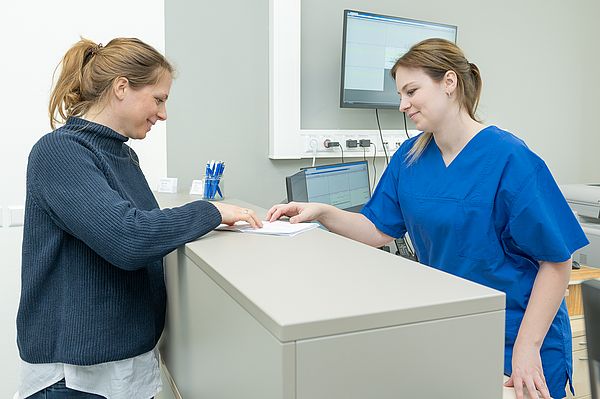 Das Bild zeigt eine Schwangere und eine Pflegekraft an der Anmeldung der Schwangerenambulanz.