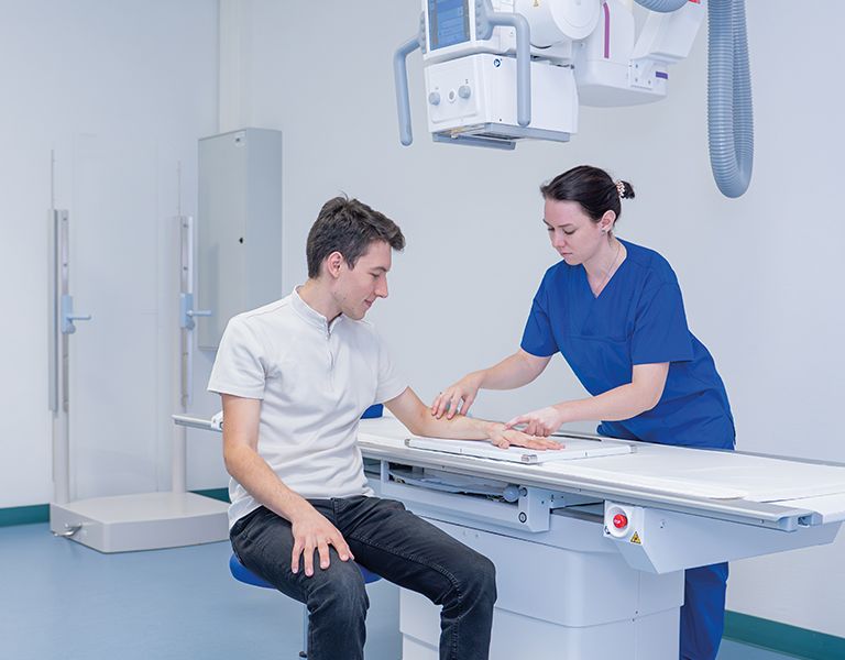 Headerbild zeigt das Röntgen einer Hand. Dabei sitzt ein Patient in der Bildmitte mit der Hand auf dem Röntgentisch, während eine MTR eine Röntgenaufnahme anfertigt.