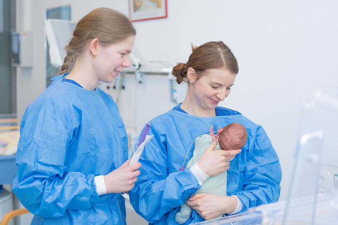 Das Bild zeigt zwei Krankenpflegerinnen mit einem Neugeborenen auf dem Arm.