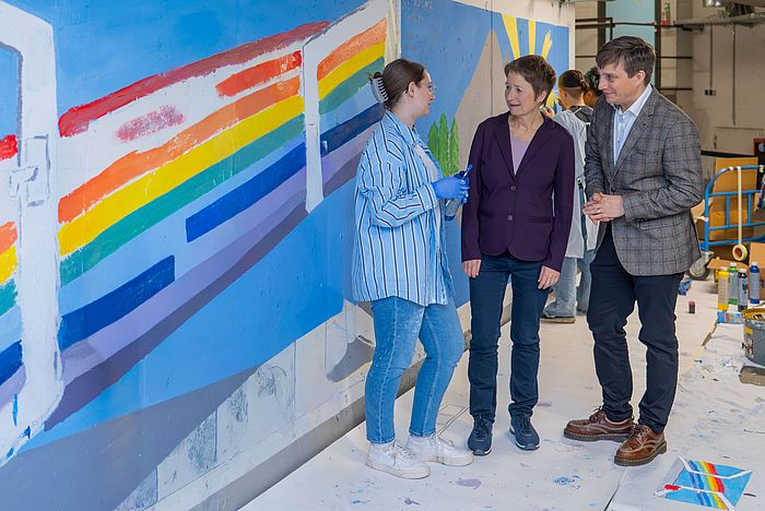 Das Bild zeigt Luisa Lehn, Lehrerin an der Drais-Gemeinschaftsschule, mit Erster Bürgermeisterin Bettina Lisbach und Prof. Dr. Dr. Martin Holderried (v.l.) vor den neu bemalten Wänden.