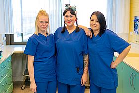 Das Bild zeigt drei Pflegekräfte in blauen Kasacks.