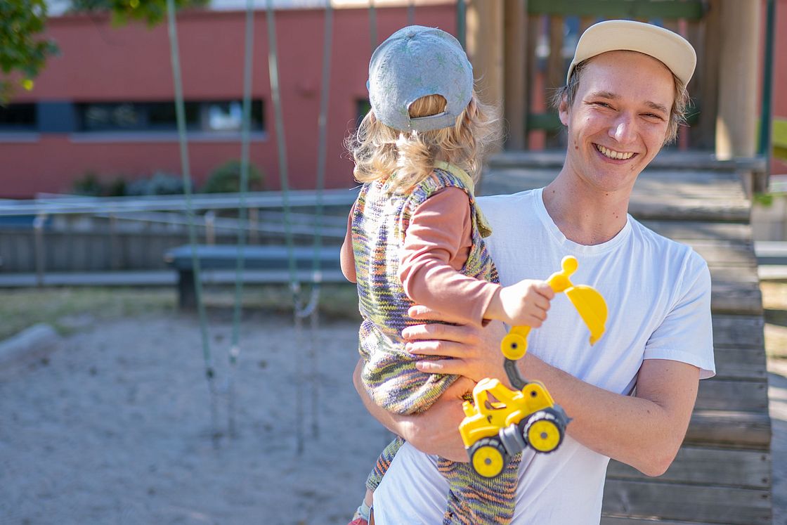 Das Bild zeigt einen Mitarbeiter, der auf einem Spielplatz sein Kind auf dem Arm hält