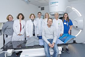 Das Bild zeigt das interdisziplinäre Team um Prof. Katja Lindel (3.v.l.), Prof. Armin Luik (4.v.l.), Dr. Antje Wark (3. v.r.) und Prof. Julian Widder (2.v.r.) mit einem Patienten (sitzend auf dem Linearbeschleuniger).