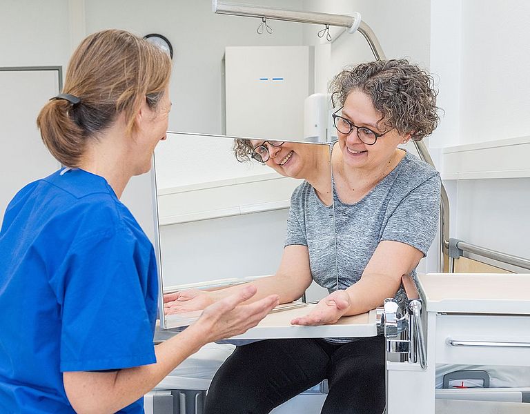 Das Bild zeigt eine Ergotherapeutin bei der Arbeit mit einer Patientin. Es wird eine Wahrnehmungsübung mit Spiegel gezeigt