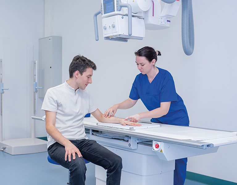 Headerbild zeigt das Röntgen einer Hand. Dabei sitzt ein Patient in der Bildmitte mit der Hand auf dem Röntgentisch, während eine MTR eine Röntgenaufnahme anfertigt.